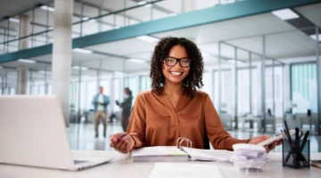 Eine lächelnde Geschäftsfrau sitzt an ihrem Schreibtisch in einem modernen Büro, hält Dokumente und schaut mit Brille in die Kamera. Vor ihr steht ein Laptop. Im Hintergrund sind Glaswände und Menschen in Bewegung zu sehen.