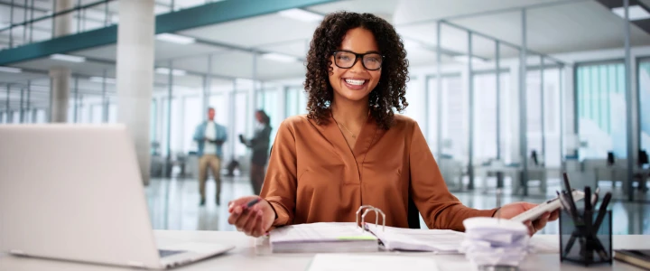 Eine lächelnde Geschäftsfrau sitzt an ihrem Schreibtisch in einem modernen Büro, hält Dokumente und schaut mit Brille in die Kamera. Vor ihr steht ein Laptop. Im Hintergrund sind Glaswände und Menschen in Bewegung zu sehen.