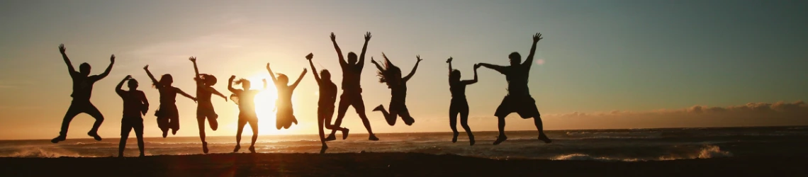 Vor einem Sonnenuntergang und dem Meer springen mehrere Menschen nebeneinander hoch in die Luft