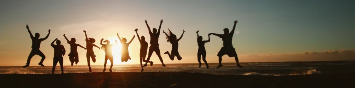 Vor einem Sonnenuntergang und dem Meer springen mehrere Menschen nebeneinander hoch in die Luft