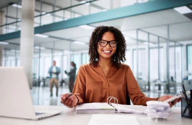 Eine lächelnde Geschäftsfrau sitzt an ihrem Schreibtisch in einem modernen Büro, hält Dokumente und schaut mit Brille in die Kamera. Vor ihr steht ein Laptop. Im Hintergrund sind Glaswände und Menschen in Bewegung zu sehen.