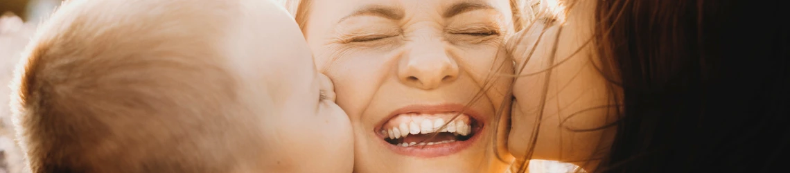 Nahaufnahme einer lachenden Mutter mit geschlossenen Augen, deren beide Kinder sie jeweils links und rechts auf die Wange küssen